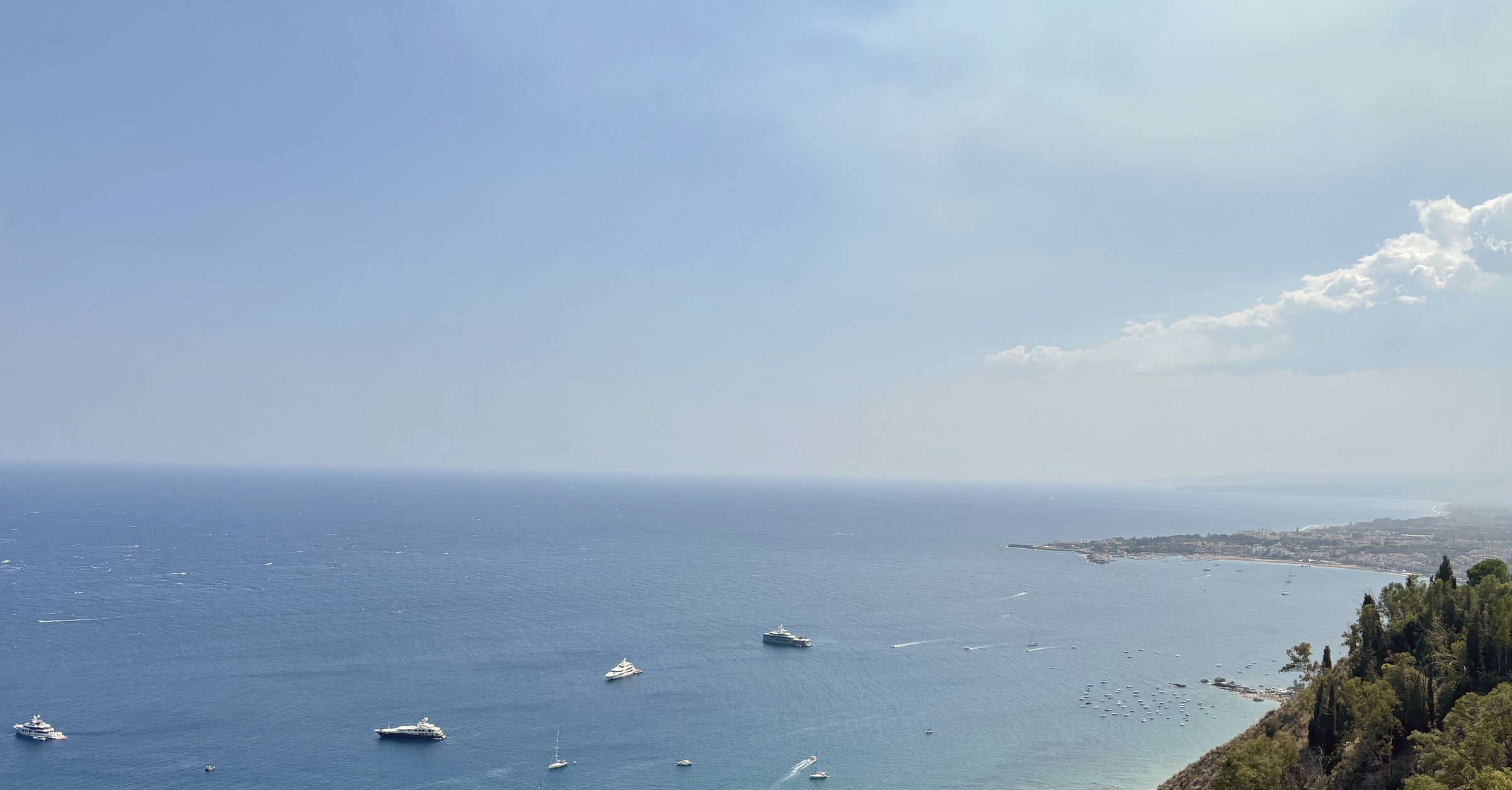 A rocky coastline curves away into a hazy blue sea, terraced hillside town above the water, seen from high on the cliffside.